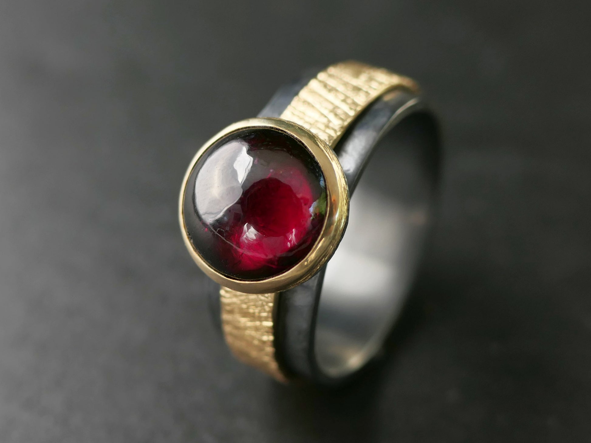 Viking-inspired bold garnet ring with a blood red cabochon set in 14k yellow gold bezel and dark oxidized sterling silver band with gold crosshatch detail