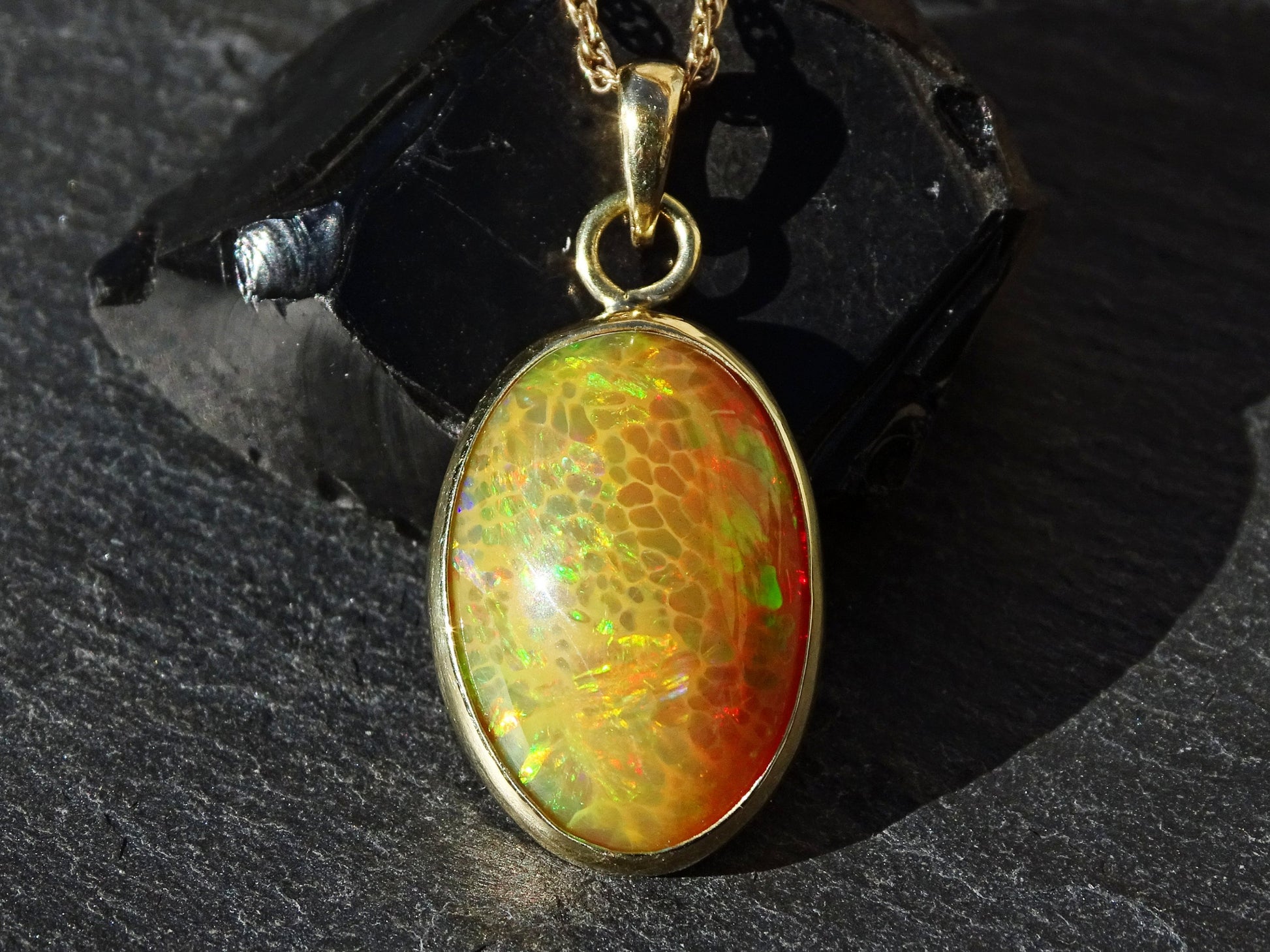 Close-up of large honeycomb welo opal pendant with vibrant red, orange, yellow, and green colors in 18k gold setting on dark stone background