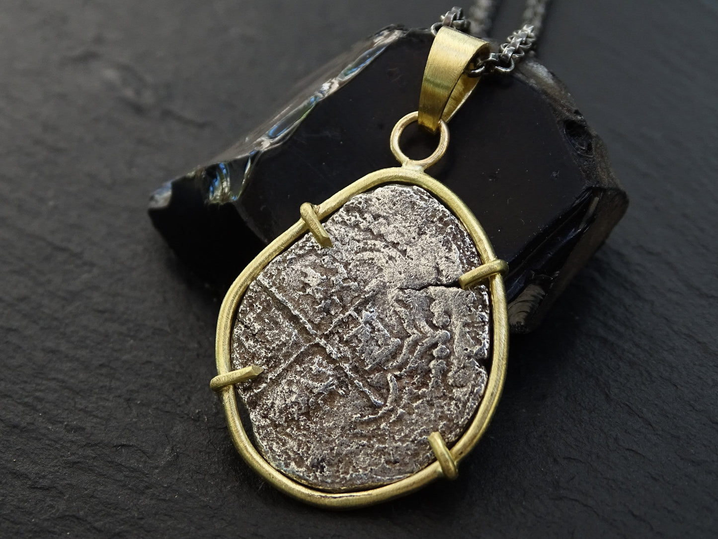 Atocha 2 Reales coin pendant set in 18k gold with black sterling silver chain on dark background