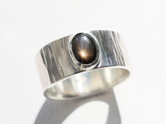 Wide silver band men's ring with black star sapphire gemstone, featuring a hammered texture resembling a waterfall or tree bark.