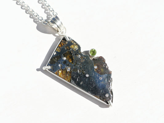 Genuine Pallasite Meteorite Necklace with a dark metallic meteorite slice, golden inclusions, silver setting, green gem and diamond accents.